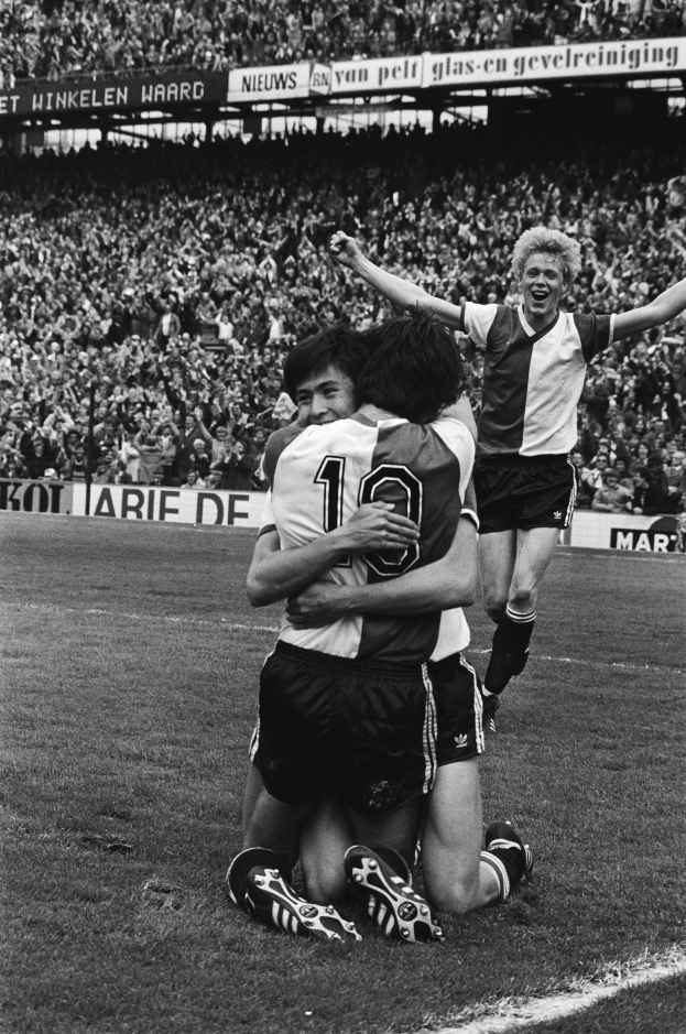 Drei Fußballspieler feiern ein Tor, indem sie sich auf dem Boden umarmen, vor einem jubelnden Publikum in einer schwarz-weißen Stadion-Szene.