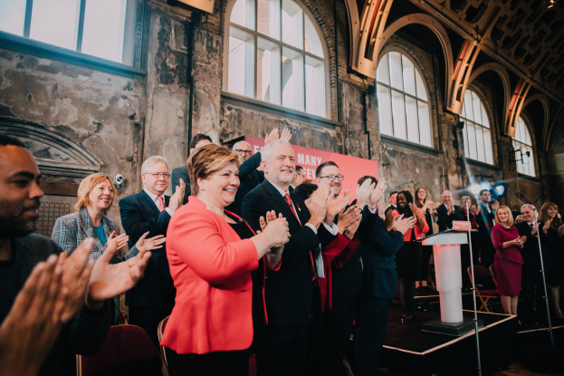 Eine Gruppe von Menschen klatscht feierlich vor einer Menge, mit einem Podium und einem Mikrofon auf der rechten Seite und einem Banner mit der Aufschrift "Covid-19 New York City Mayor Bill de Blasio" im Hintergrund.