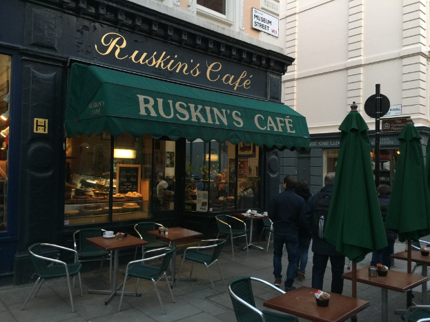Eine Gruppe von Menschen steht vor einem Café mit einem Schild, auf dem "Ruskin's Café" steht.
