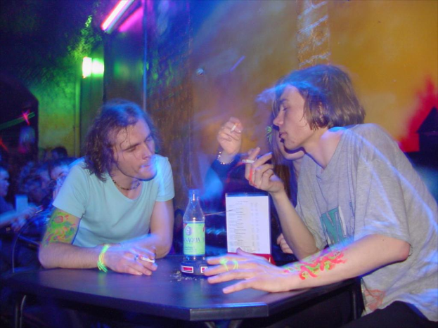 Ein Tisch mit einer Flasche, einer Speisekarte und einem Aschenbecher in der Mitte einer Indoor-Szene, mit zwei Männern und einer Frau, die Zigaretten rauchen.