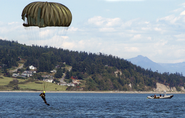 Eine Person gleitschirmfliegt über einen See mit einem Boot auf der rechten Seite, Bäumen, Gebäuden, Hügeln und einem klaren blauen Himmel im Hintergrund.