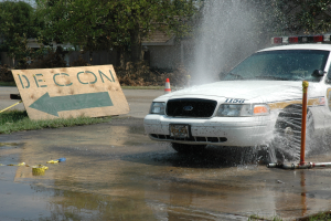 Polizeiauto fährt durch Pfütze mit verstreuten Gegenständen, darunter eine Platte, eine Verkehrsinsel, Bäume, Gebäude und einen klaren blauen Himmel im Hintergrund.
