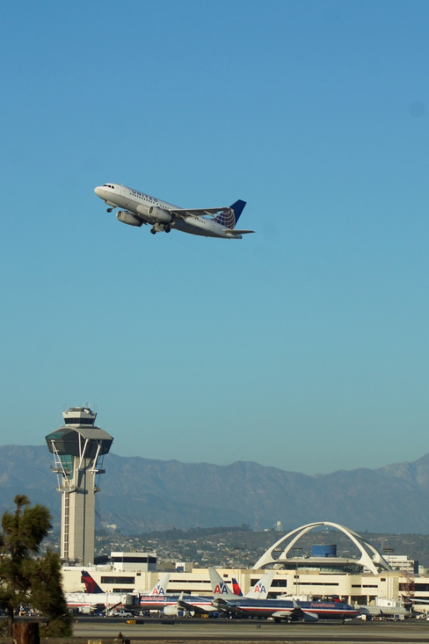 Eine Flughafenansicht mit einem weißen Kontrollturm, geparkten Flugzeugen, entfernten Gebäuden und einem Flugzeug am Himmel.