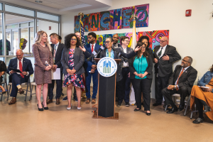 Eine Gruppe von Menschen vor einem Podium mit einem Mikrofon und einer Tafel mit der Aufschrift "Sancho-Stadtrat tritt dem Stadtrat bei", mit anderen im Hintergrund sitzend und stehend, Flaggen, eine Tafel, einen Feuerlöscher und Deckenleuchten an der Rückwand.