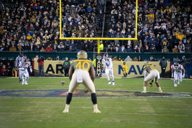 Fussballspieler mit Helmen und Sportbekleidung konkurrieren auf einem Feld, mit Zuschauern auf den Rängen und Stadionwerbung im Hintergrund.