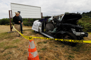 Ein Polizeibeamter in Uniform steht auf dem Gras neben einem schwer beschädigten Auto mit eingedellter Haube und zersplitterten Fenstern, mit einem Verkehrskegel und einem Band an einem Pfahl in der Nähe und Bäumen und einem bewölktem Himmel im Hintergrund.