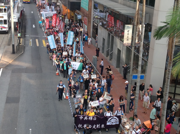 Eine große Gruppe von Menschen marschiert auf einer Straße in Hong Kong, mit Schildern und Plakaten in den Händen, mit Bäumen, Glasfassaden, Fahrzeugen und Schildern auf beiden Seiten.
