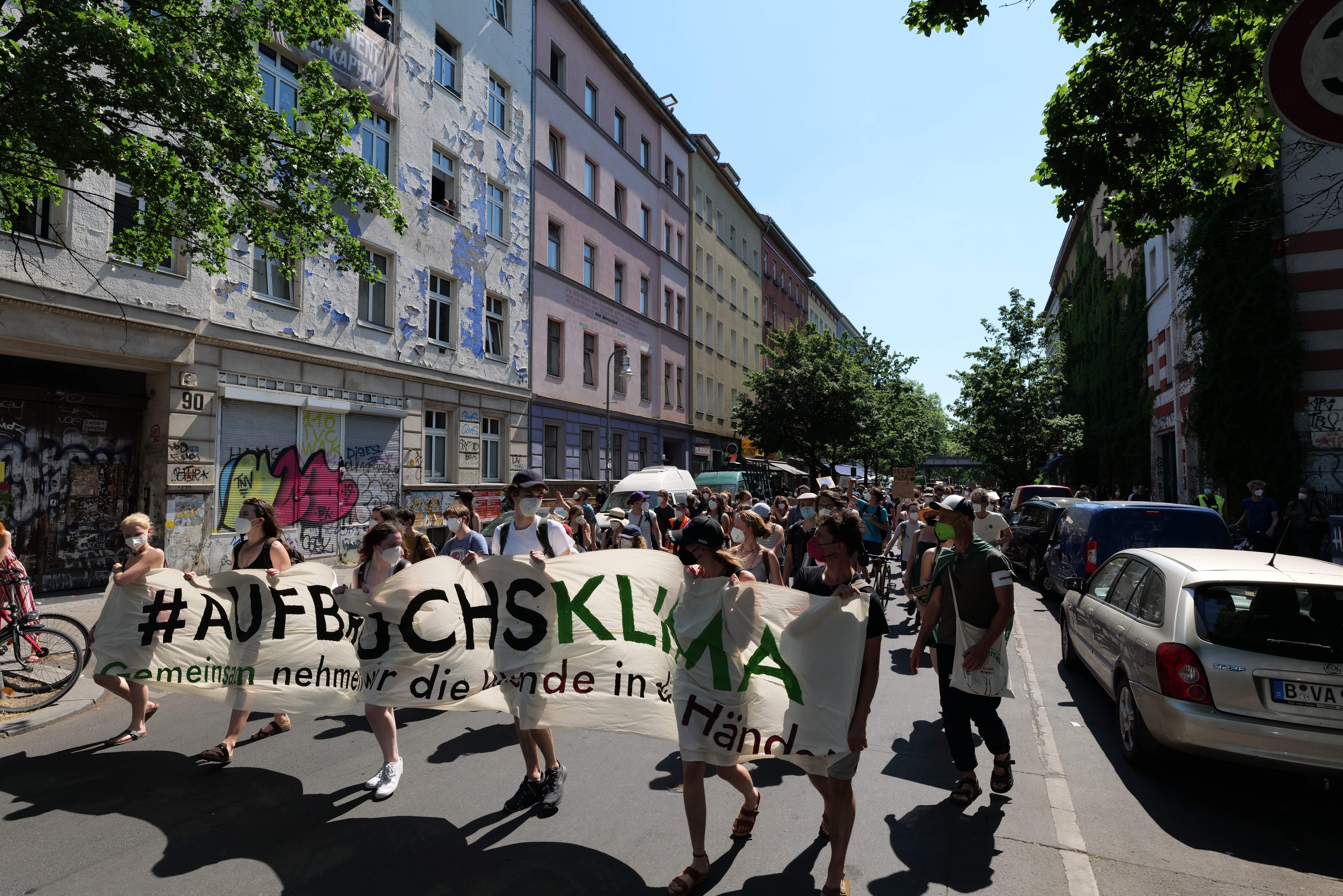 Eine Gruppe von Menschen marschiert mit einem Transparent die Straße entlang, mit parkenden Autos auf der rechten Seite und einem Radfahrer auf der linken Seite, vor Gebäuden und Bäumen in Berlin.