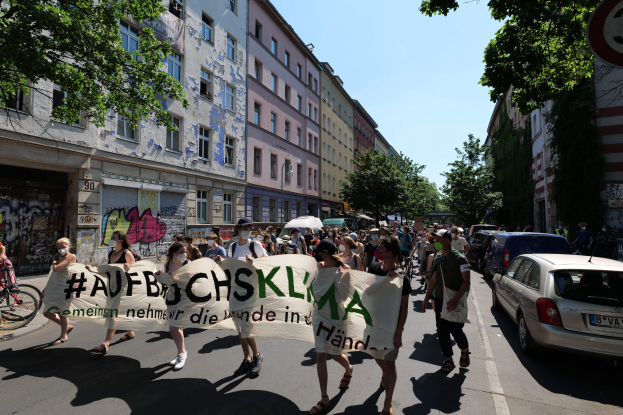 Eine Gruppe von Menschen marschiert mit einem Transparent die Straße entlang, mit parkenden Autos auf der rechten Seite und einem Radfahrer auf der linken Seite, vor Gebäuden und Bäumen in Berlin.