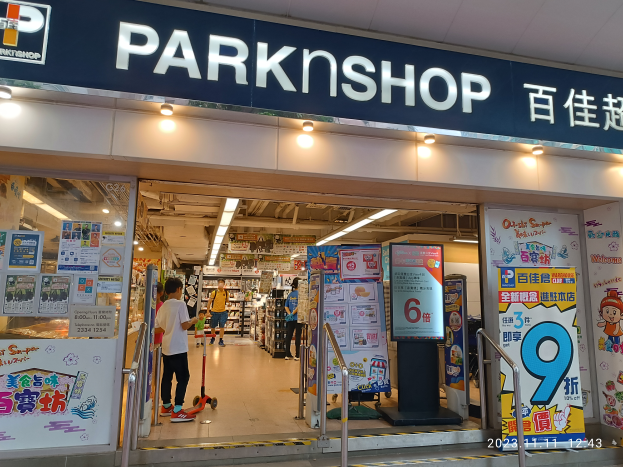 Ein Parkshop in einem Einkaufszentrum mit einem Schild, auf dem "Park Shop" steht, und einer Gruppe von Menschen auf dem Boden, umgeben von Metallstangen, Schildern, Bannern, Gestellen mit Gegenständen und Deckenleuchten.