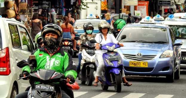 Eine Gruppe von Menschen auf Motorrädern fährt eine belebte Straße entlang mit Autos und anderen Fahrzeugen, einige Fahrer tragen Helme, Gebäude, Bäume, Schilder mit Text und Geschäfte im Hintergrund.