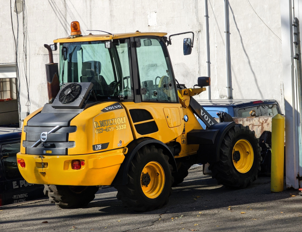 Ein gelber Radlader steht auf einer Straße vor einem Gebäude mit einer Wand im Hintergrund und anderen Fahrzeugen in der Nähe.