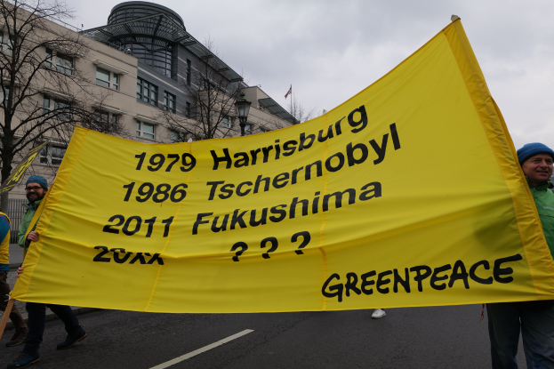 Gruppe von Menschen geht eine von Bäumen gesäumte Straße entlang und hält eine gelbe "Greenpeace"-Fahne hoch, mit Gebäuden und Himmel im Hintergrund.