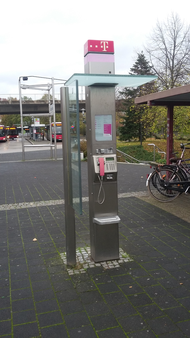 Ein Münzfernsprecher steht an einer Straße neben einer Bushaltestelle, daneben ein Fahrrad geparkt; im Hintergrund gibt es Bäume, Fahrzeuge auf der Straße, eine Brücke und einen klaren blauen Himmel.