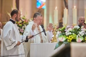 Bischof der Kirche des Heiligen Grabes in Priestergewand vor einem Podium mit Mikrofon, umgeben von Menschen, mit einem Kerzen- und Blumenaltar rechts, Blumensträußen und einem Bleiglasfenster im Hintergrund.