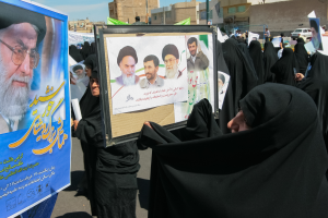 Eine Gruppe von Frauen in schwarzen Abayas hält Plakate mit Fotos von Ayatollah Ali Khamenei hoch, im Hintergrund sind Gebäude, Pfähle, Fahrzeuge und ein klarer blauer Himmel zu sehen.