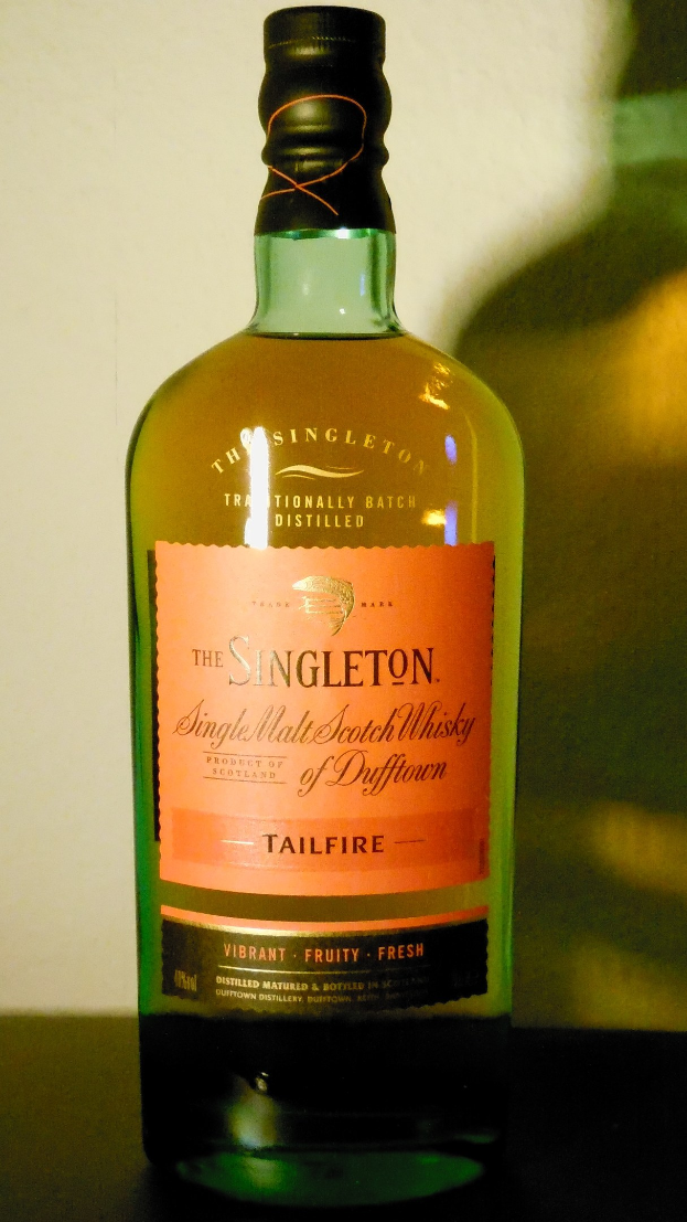 Eine tiefe bernsteinfarbene Flasche Singleton of Dufftown Tailfire Single Malt Scotch Whisky auf einem Tisch, mit einem Etikett, das schwarze Schrift und ein Wappen mit einem Löwen und einer Burg zeigt.