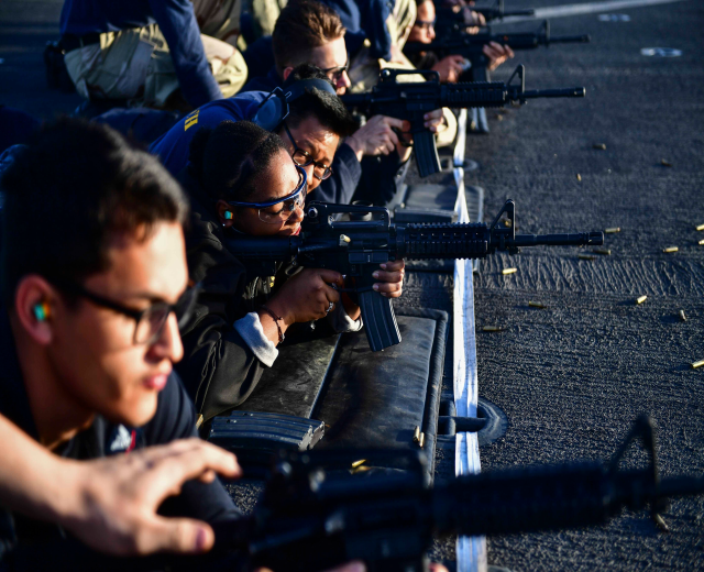 Eine Gruppe von Menschen, die auf dem Boden sitzen und jeweils ein Gewehr halten, mit Patronen, die vor ihnen auf der Straße verstreut sind, wahrscheinlich an einem Trainingsübung teilnehmen.