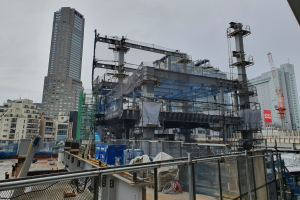 Großer Baustelle in einer Stadt für ein neues Kraftwerk, umgeben von einem Metallzaun, mit Gebäuden, Masten, einer Fahne, Fahrzeugen, Drähten und einem bewölktem Himmel.