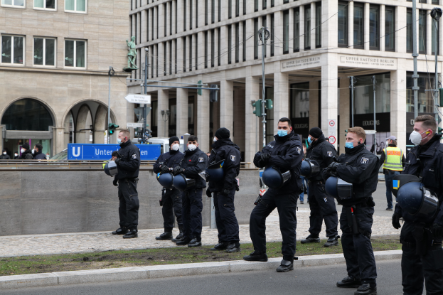 Eine Gruppe von Polizisten in schwarzen Uniformen und Masken steht vor einem Gebäude mit Glasfenstern und Säulen in Berlin, Deutschland, einige halten Helme, mit Laternen, Verkehrsampeln, Texttafeln, einer Statue, anderen Menschen und grasbewachsenem Boden im Hintergrund.