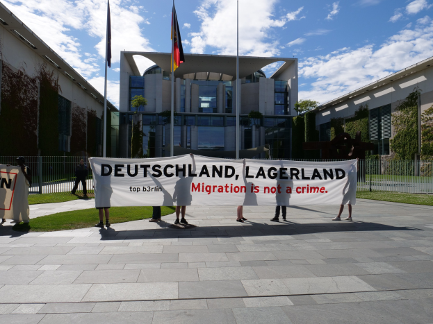 Gruppe von Menschen mit einem Transparent mit der Aufschrift "Deutschland, Lagerland Migration ist kein Verbrechen" vor einem Fensterbau mit einer Metallumzäunung, umgeben von Gras, Pflanzen, Bäumen und Fahnenmästen unter einem bewölkten Himmel.