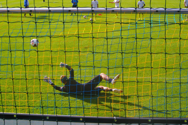 Ein Fußballspieler macht eine Parade vor einem Tor, mit einem Netz im Vordergrund und einigen Menschen im Hintergrund, während mehrere Bälle auf dem Boden verstreut sind.