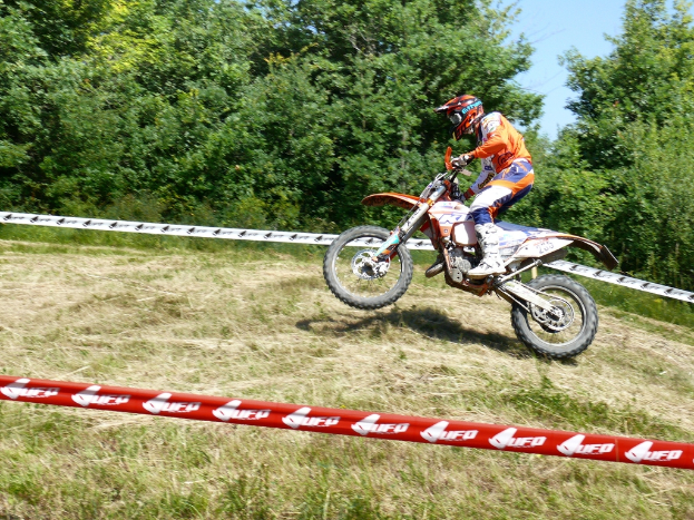Eine Person fährt ein Fahrrad mit Helm, umgeben von Absperrband, mit Bäumen, Gras und Himmel im Hintergrund.