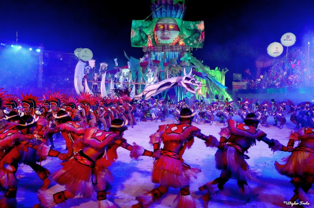 Eine Gruppe von Menschen in bunten Kostümen tanzt auf einer Bühne vor einem sitzenden Publikum, mit Lichtern, Polen, Ballons und einem Bildschirm, der eine Person in der Hintergrund zeigt, unter einem sichtbaren Himmel, bei der Rio-Karneval 2014.