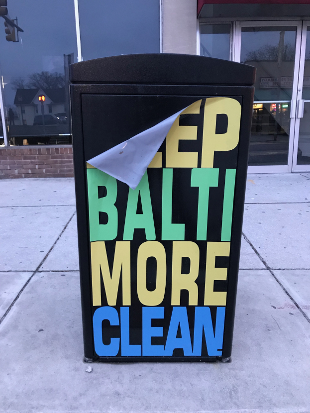 Ein schwarzer Müllcontainer mit der Aufschrift "Keep Baltimore More Clean" vor einem Gebäude mit Glastüren