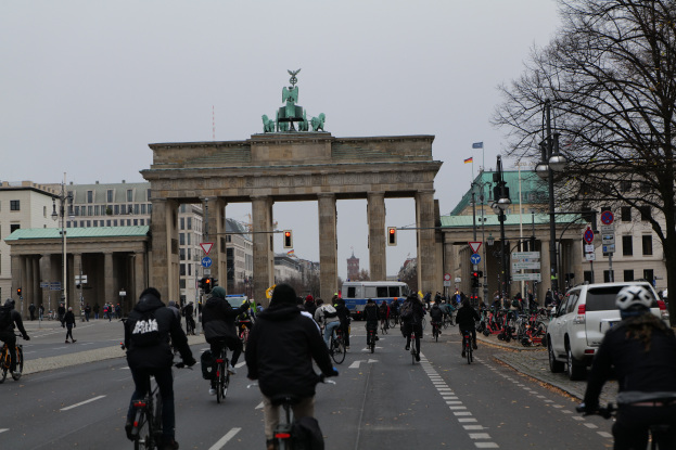 Eine Gruppe von Menschen, die Fahrräder auf einer Straße vor dem Brandenburger Tor in Berlin, Deutschland, fährt, mit Bäumen, Laternen, Verkehrszeichen, Schildern und Gebäuden mit Fenstern im Hintergrund und dem Himmel in der Ferne.