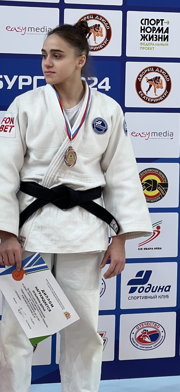 Frau in weißem Judo-Anzug mit schwarzem Gürtel auf einem Podest, die eine Medaille und ein Zertifikat hält, mit einem Logo-Plakat im Hintergrund.