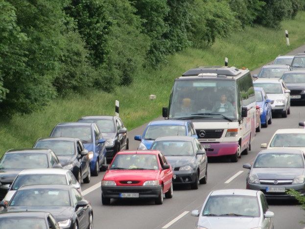 Ein Stau auf einer Autobahn mit mehreren Autos und einem Van, Menschen sichtbar in den Fahrzeugen, und Bäume und Gras im Hintergrund.