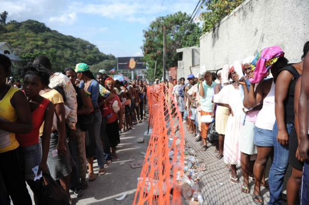 Lange Schlange von Menschen, die an einer Wahllokalwarten, mit einem Zaun, einer Wand, Bäumen, Gebäuden, Strommasten, Drähten, einer Werbetafel und Himmel im Hintergrund.