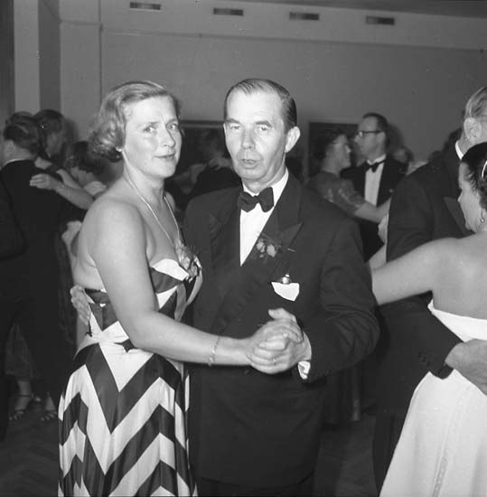 Ein Mann in einem schwarzen Blazer, weißem Hemd und schwarzen Hosen tanzt mit einer Frau in unbekannter Kleidung auf einer formellen Veranstaltung, indem sie sich an den Händen halten und zur Musik schwenken, vor einer Wand als Hintergrund.