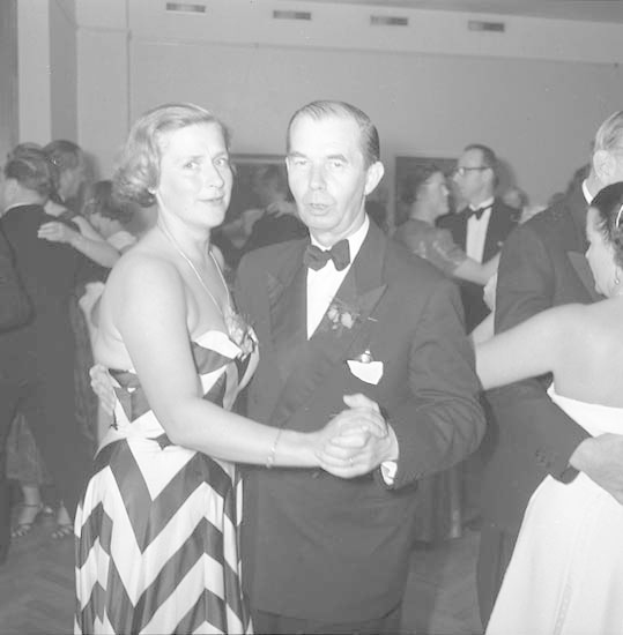 Ein Mann in einem schwarzen Blazer, weißem Hemd und schwarzen Hosen tanzt mit einer Frau in unbekannter Kleidung auf einer formellen Veranstaltung, indem sie sich an den Händen halten und zur Musik schwenken, vor einer Wand als Hintergrund.