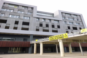 Ein großes Gebäude mit einem "Flüchtlinge Willkommen"-Schild, umgeben von Säulen und einem Geländer, unter einem Himmel-Hintergrund.