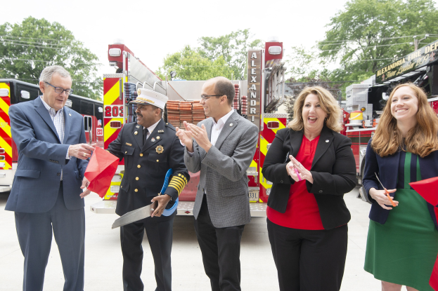 Gruppe von Menschen, die bei einer Einweihungszeremonie der Alexandria Fire Department klatschen und lächeln, mit zwei Personen, die eine Schere und ein rotes Band halten, vor einem Feuerwehrfahrzeug.