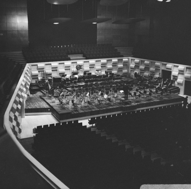 Ein Schwarz-Weiß-Foto eines großen Auditoriums mit einer Bühne in der Mitte, umgeben von Stühlen, auf denen Musiker spielen.