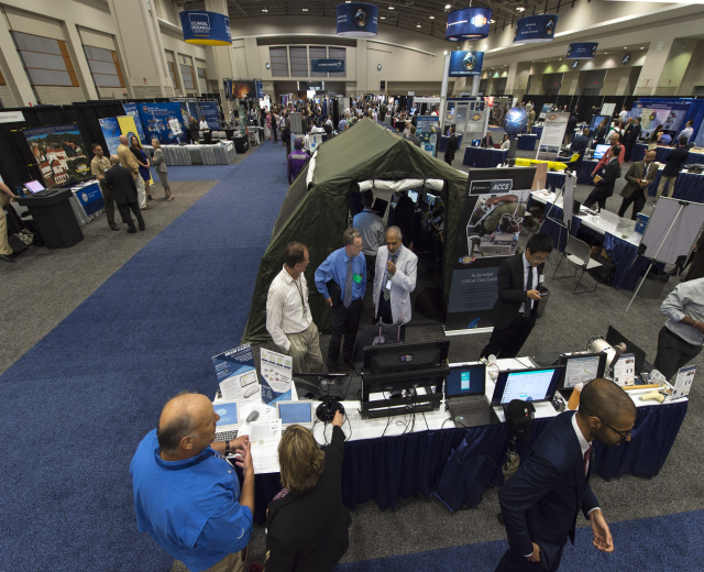 Eine große Gruppe von Menschen auf einer Karriere-Messe sitzt an Tischen mit Laptops, Monitoren und Papieren, mit einem Zelt, Schautafeln, Deckenleuchten und Fenstern im Hintergrund.
