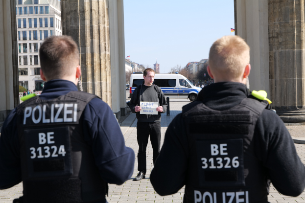 Drei Polizeibeamte stehen vor einem Gebäude mit einem Mann, der ein Papier zwischen ihnen hält, flankiert von Säulen, während Fahrzeuge, Menschen und Gebäude im Hintergrund zu sehen sind.