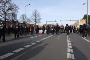 Gruppe von Polizisten in schwarzen Uniformen und Helmen auf der Seite einer Straße mit Laternenmasten, Ampeln, Bäumen, Gebäuden und einem klaren blauen Himmel im Hintergrund.