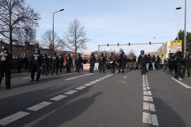 Gruppe von Polizisten in schwarzen Uniformen und Helmen auf der Seite einer Straße mit Laternenmasten, Ampeln, Bäumen, Gebäuden und einem klaren blauen Himmel im Hintergrund.