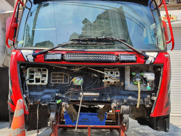 Ein roter Lkw steht vor einem Geb\c3\a4ude, mit einem Verkehrskegel links und einem Rollladen rechts, w\c3\a4hrden mit Text und Zahlen im Hintergrund sichtbar sind.