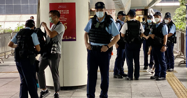 Polizeibeamte in Uniform mit Mund-Nasen-Schutz stehen vor einem Gebäude, einige halten Kameras, mit einem Banner, Geländern, Pflanzen, Bäumen und Deckenleuchten im Hintergrund.