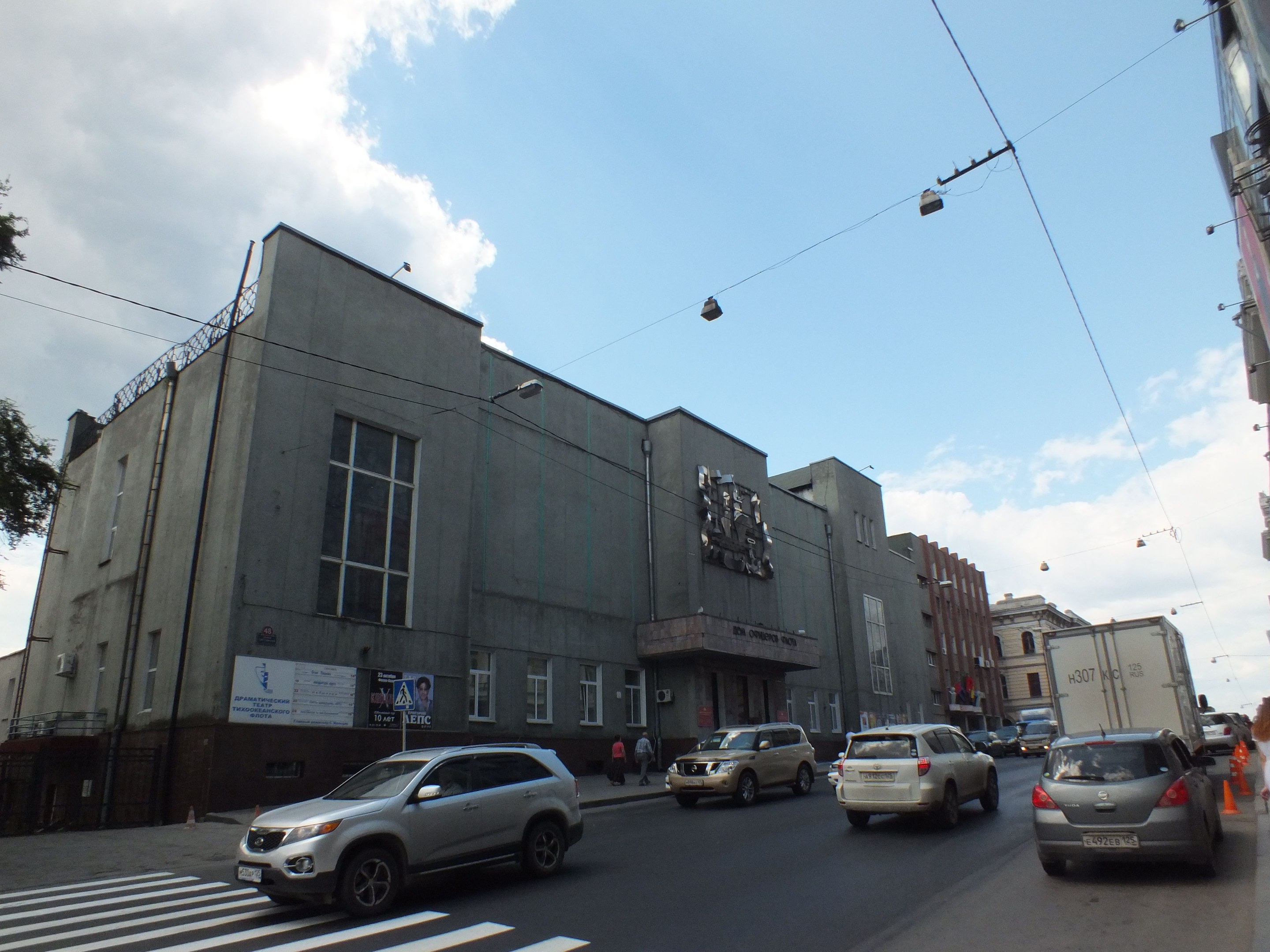 Stadtstra├če mit fahrenden Autos, eine Zebrüberquerung, Verkehrskegel, Fu├čg├Ąnger auf dem Gehweg, Geb├Ąude mit Fenstern, Laternen, Strommasten mit Kabeln, B├Ąume und ein bew├Âlktes Himmel.
