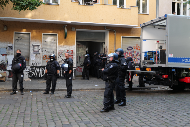 Eine Gruppe von Polizisten in Helmen und mit Waffen steht vor einem Gebäude mit Fenstern, Rollläden und Graffiti, daneben parkt ein Lastwagen und es gibt Bäume im Hintergrund.