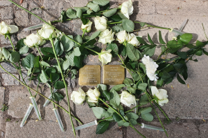 Ein Kränzchen aus weißen Rosen und zwei goldfarbene, beschriftete Gegenstände auf dem Boden vor einem Berliner Denkmal.