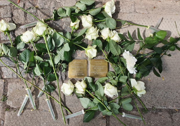 Ein Kränzchen aus weißen Rosen und zwei goldfarbene, beschriftete Gegenstände auf dem Boden vor einem Berliner Denkmal.