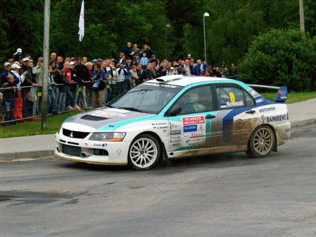 Ein Rallyeauto mit Text und Logos fährt an einer Menge vorbei, die Kameras halten, mit Geländern, Polen mit Fahnen und Bäumen im Hintergrund.