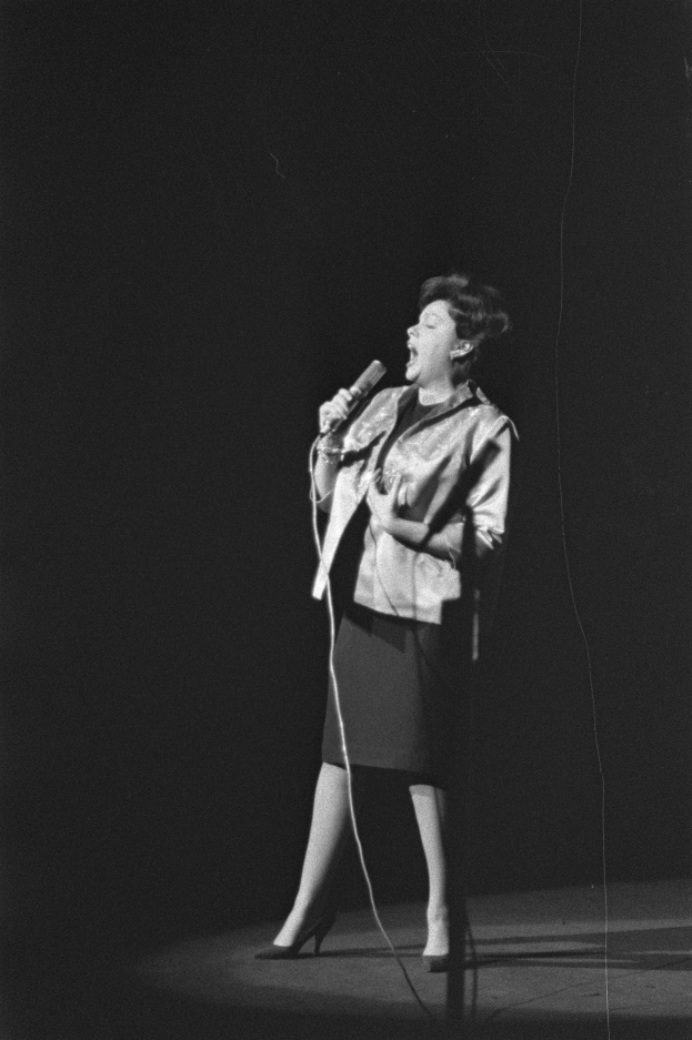 Eine Frau in einem schwarzen und weißen Outfit aus den 1950er-Jahren singt fröhlich auf einer von einem Scheinwerfer beleuchteten Bühne in ein Mikrofon.